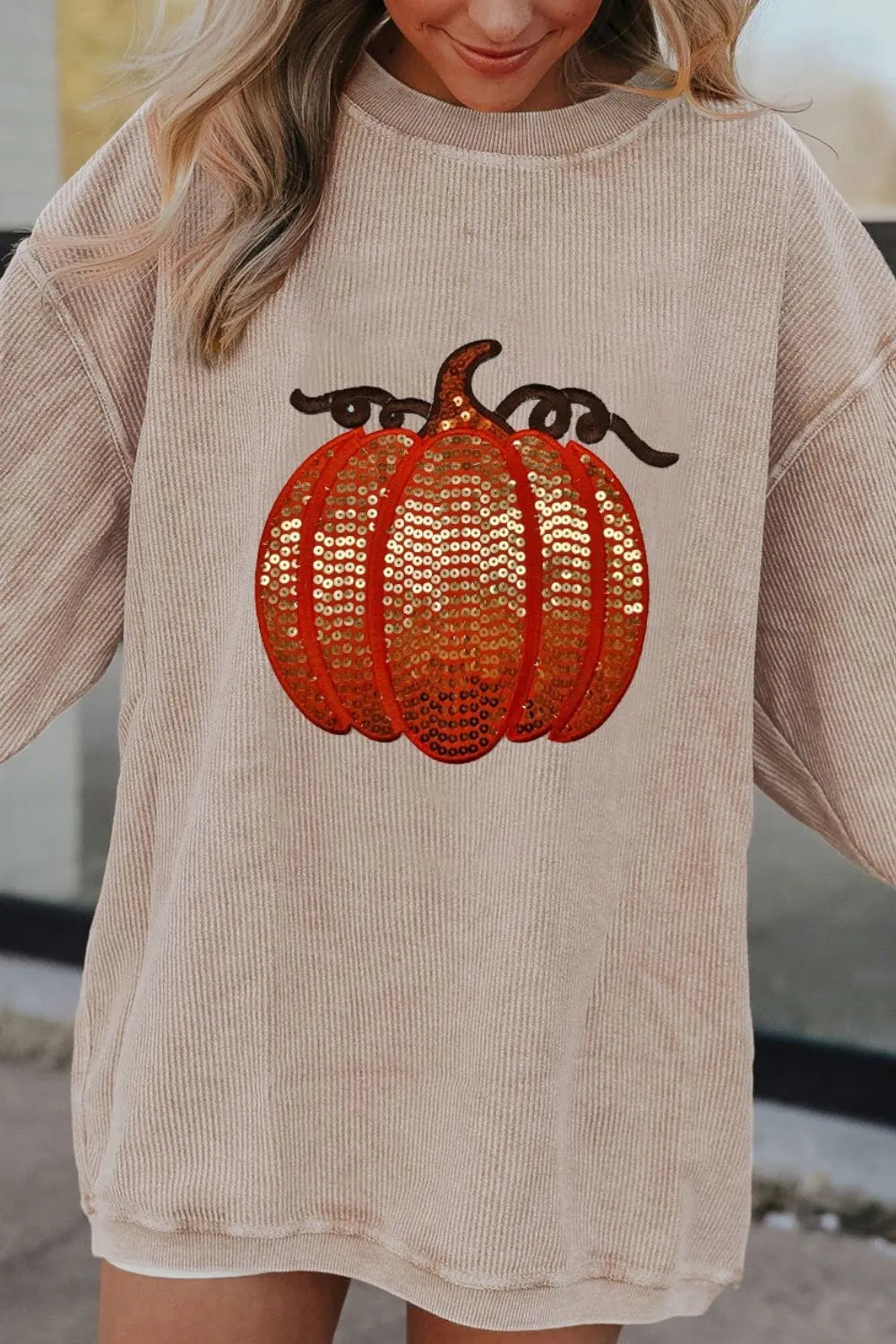 A woman wears a beige long-sleeve sweatshirt with a sequin pumpkin design.