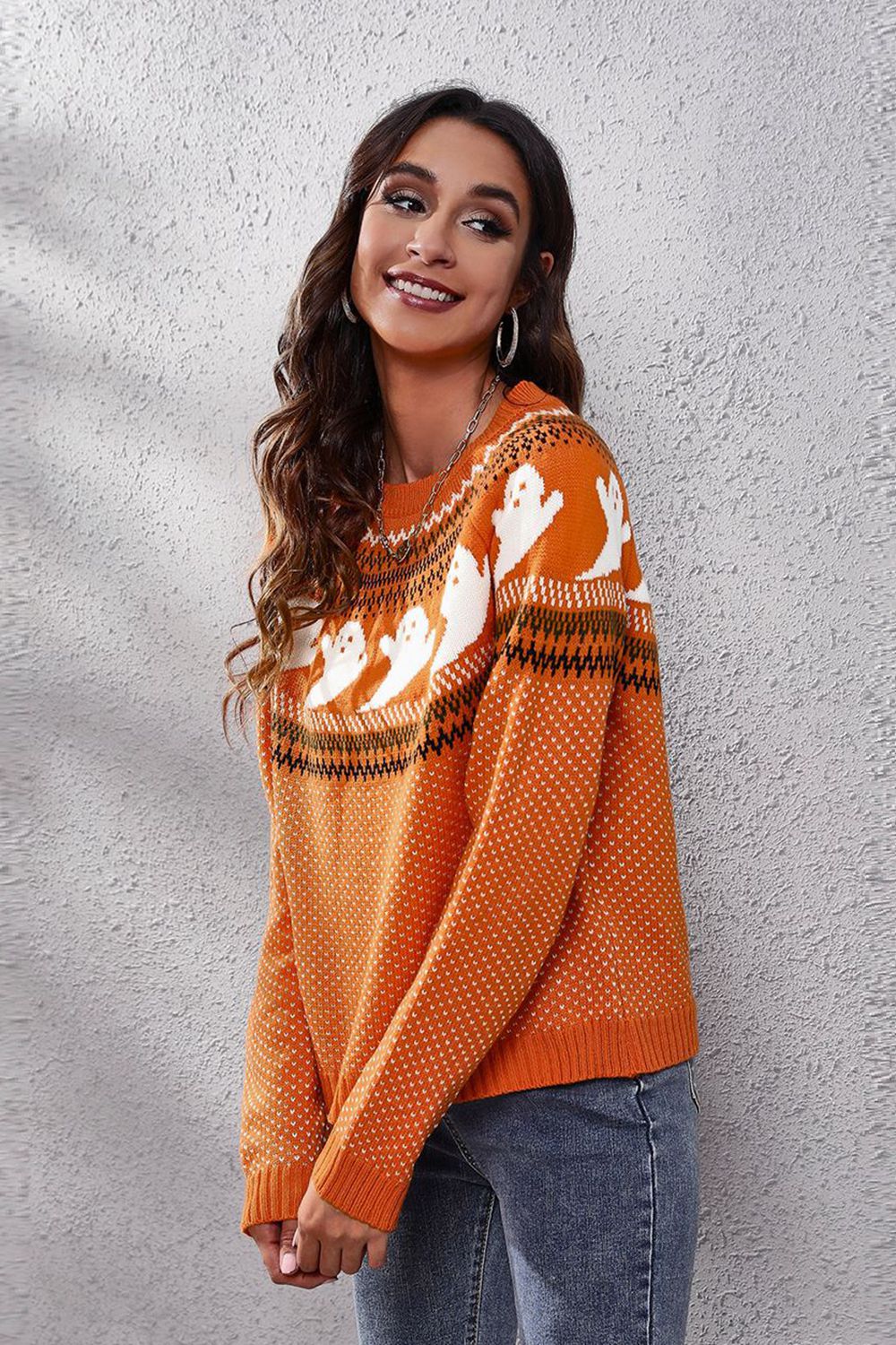 A woman smiles while wearing an orange ghost pattern round neck long sleeve sweater.