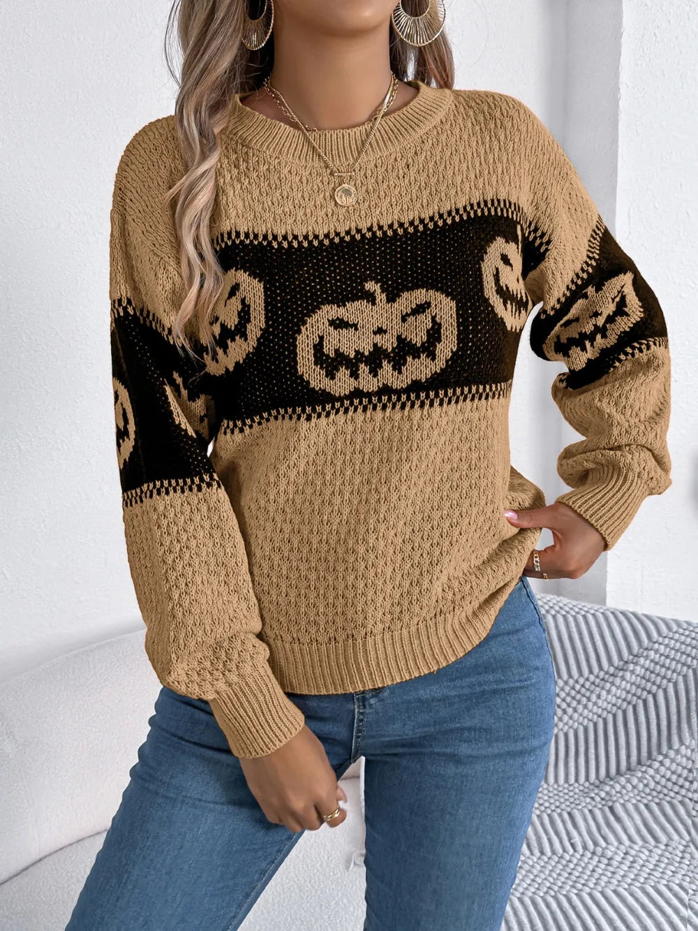 A woman wears a tan and black pumpkin-patterned long-sleeve sweater with blue jeans.