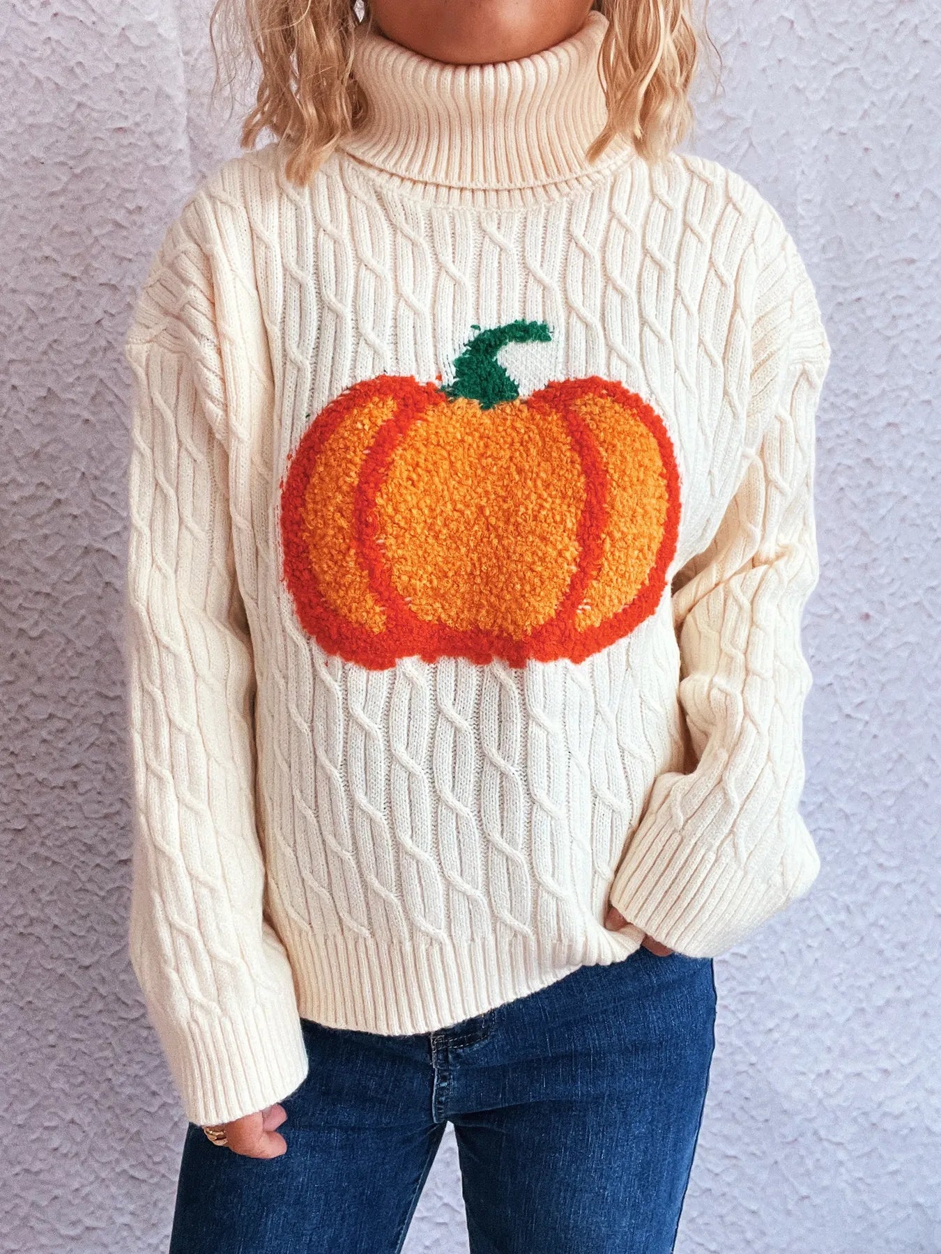 A cream cable-knit turtleneck sweater with a large orange pumpkin applique.