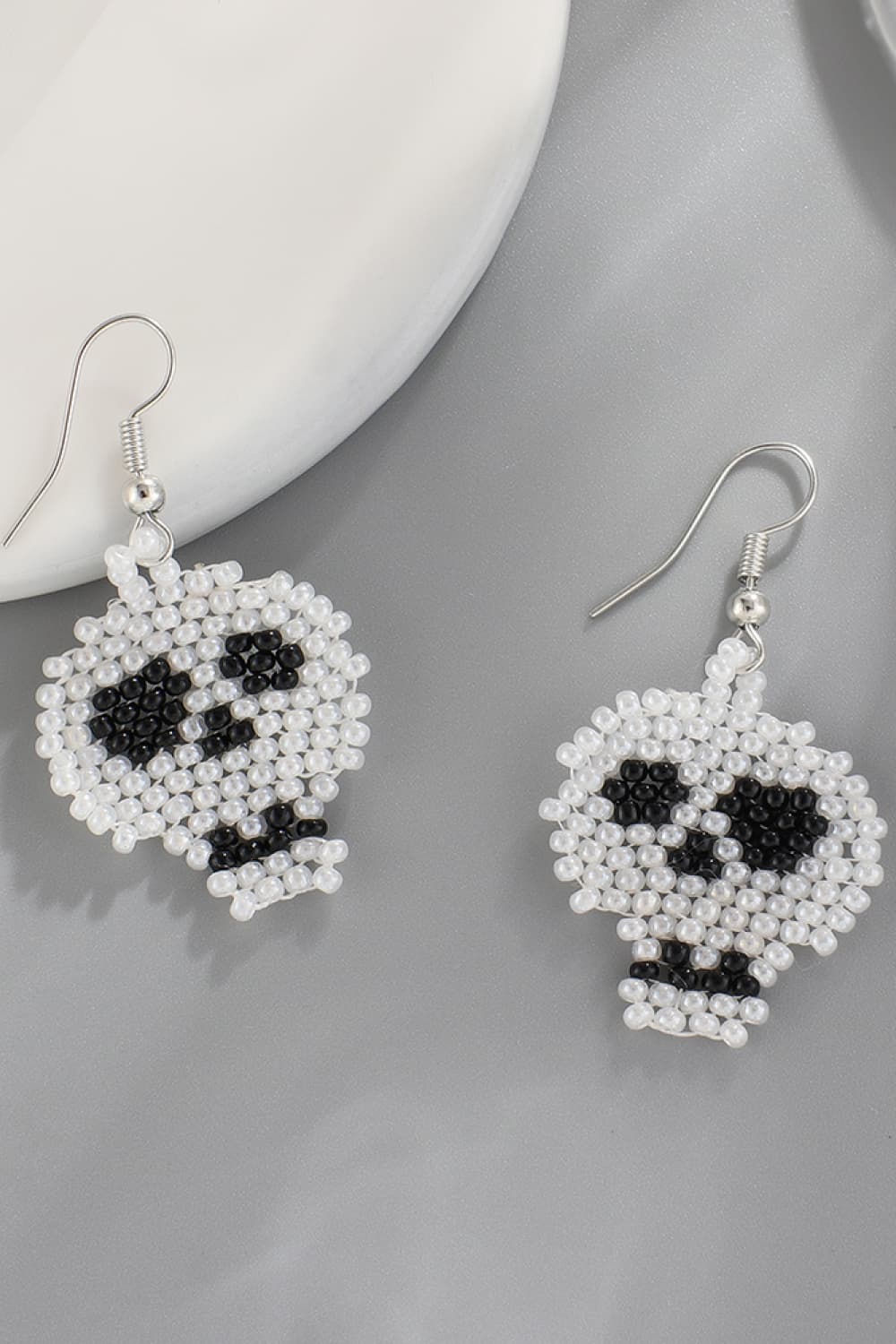 Two white and black beaded skull earrings are displayed on a gray surface.