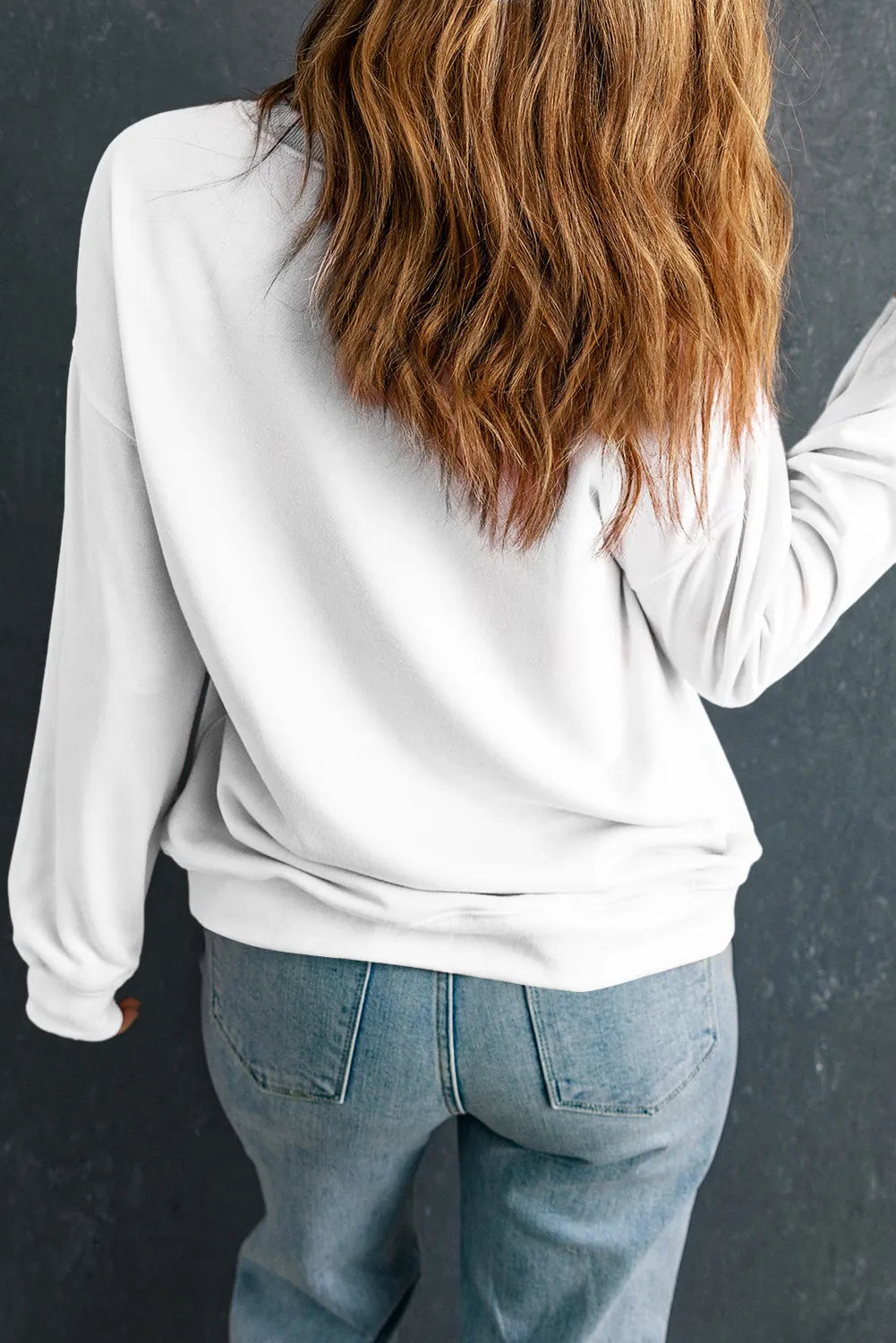 A woman from behind wears a white long-sleeve sweatshirt and blue jeans.