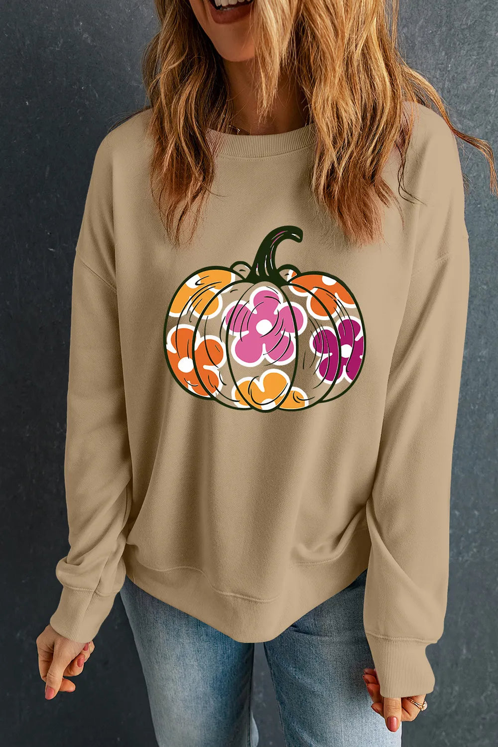 A woman wears a tan long-sleeve sweatshirt with a colorful pumpkin and flower graphic.