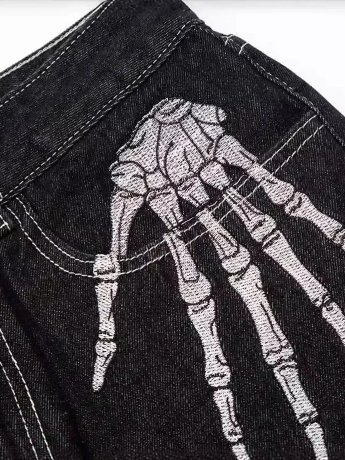 Black denim jeans with a white embroidered skeleton hand on the upper thigh.