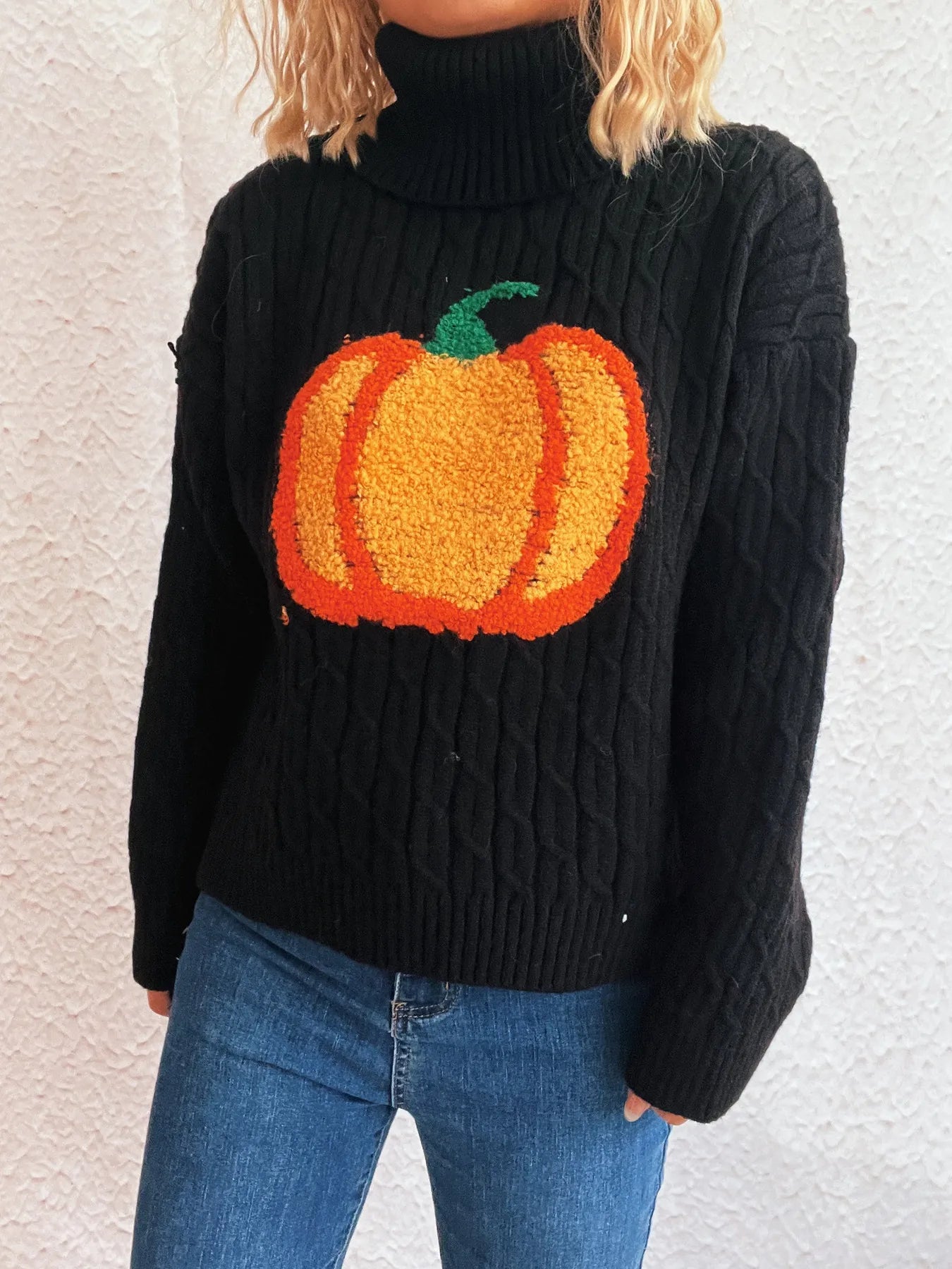 Black cable-knit turtleneck sweater with an orange pumpkin applique, worn with blue jeans.