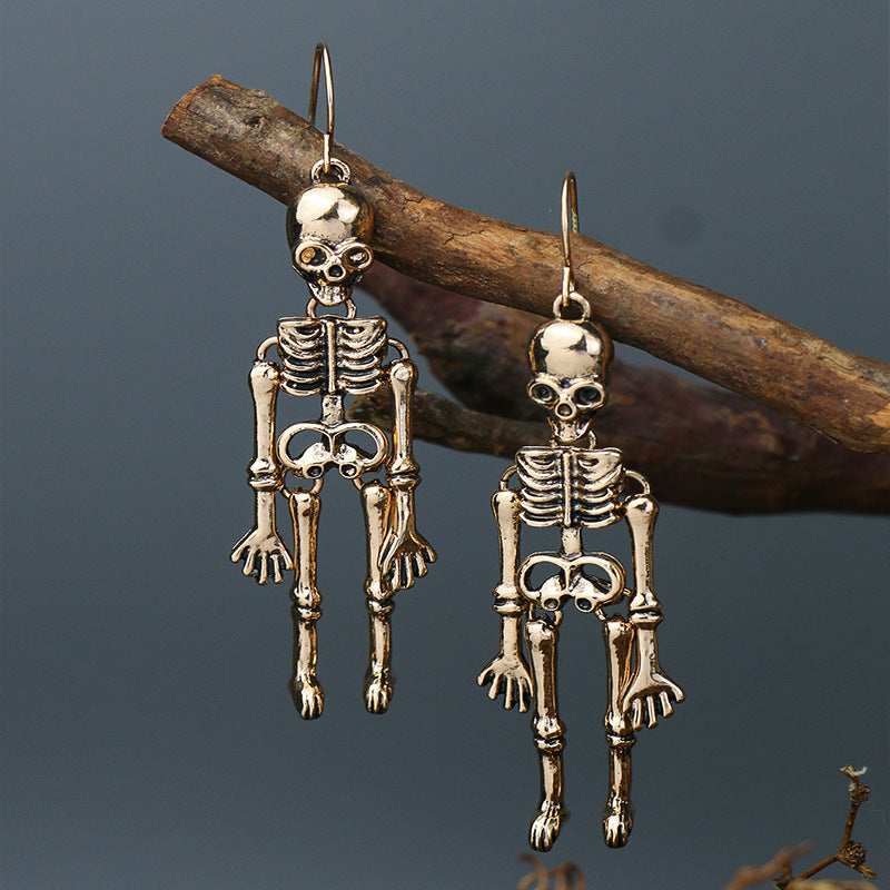 Two gold-colored skeleton alloy earrings hang from a tree branch against a dark background.