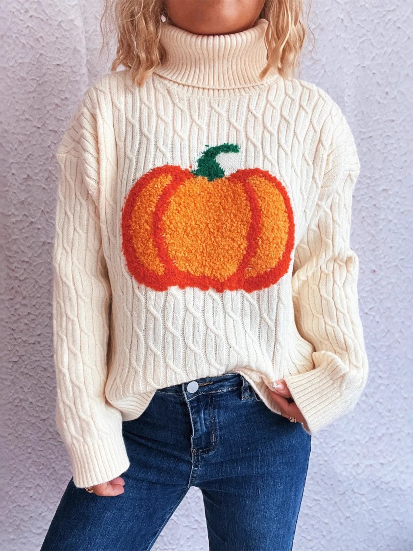 A woman wears a cream cable-knit turtleneck sweater with an orange pumpkin applique.