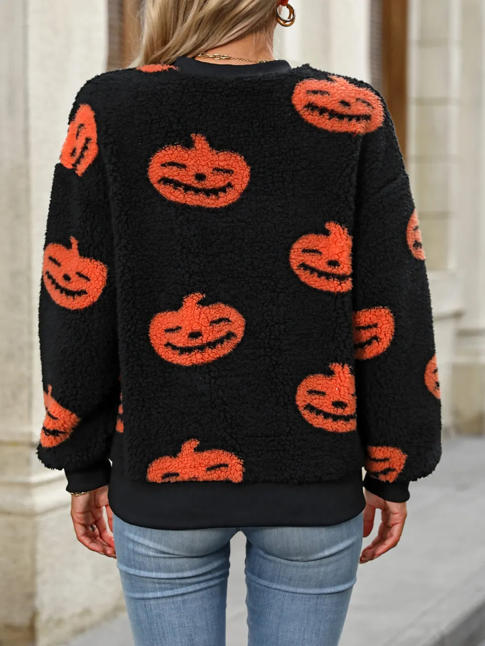 A woman from behind wears a black fuzzy sweater with orange pumpkin faces.