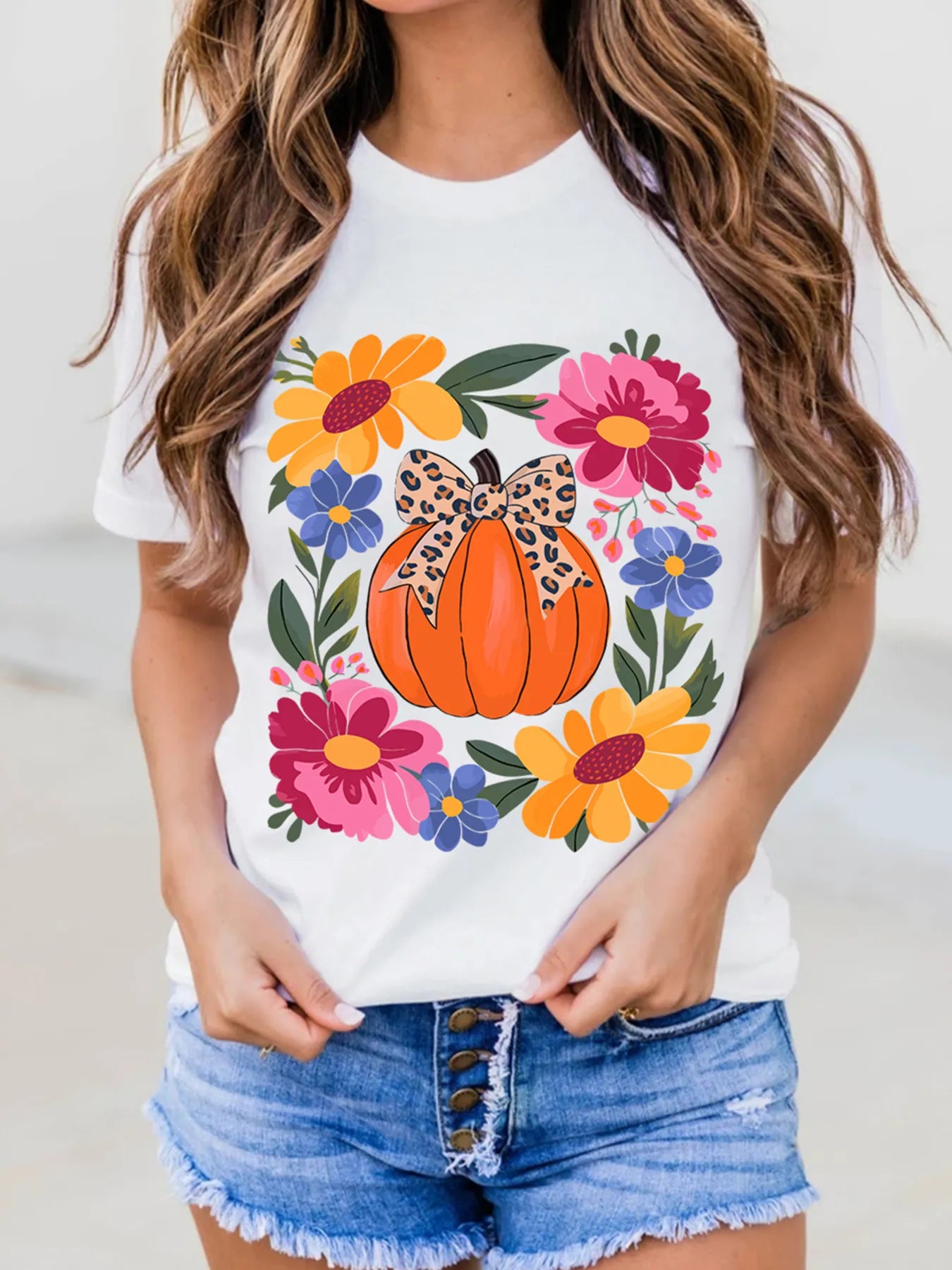 A woman wears a white t-shirt with a pumpkin and floral graphic, and denim shorts.