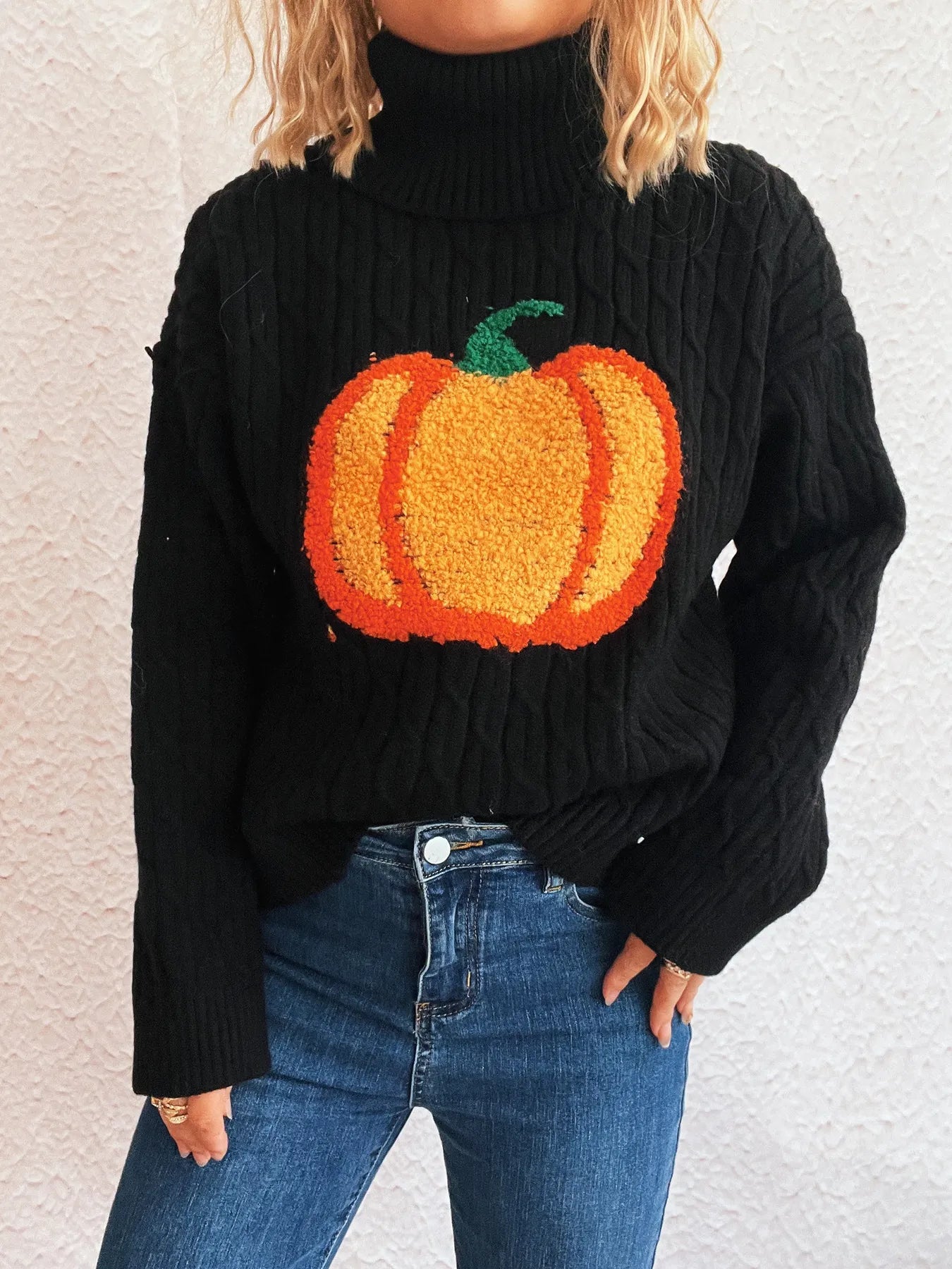 Black cable-knit turtleneck sweater with an orange pumpkin applique, paired with blue jeans.