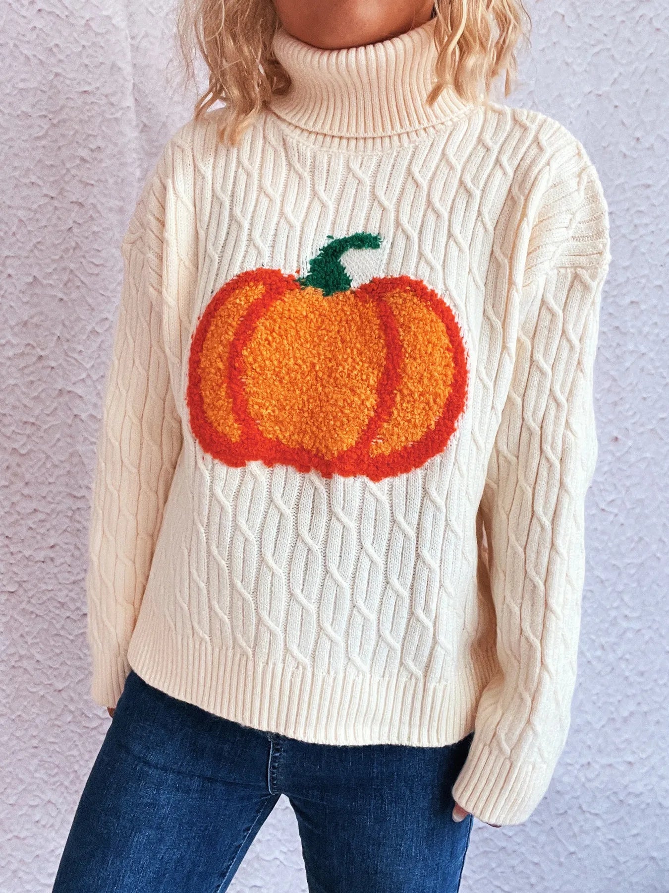 A cream cable-knit turtleneck sweater with a large orange pumpkin applique.