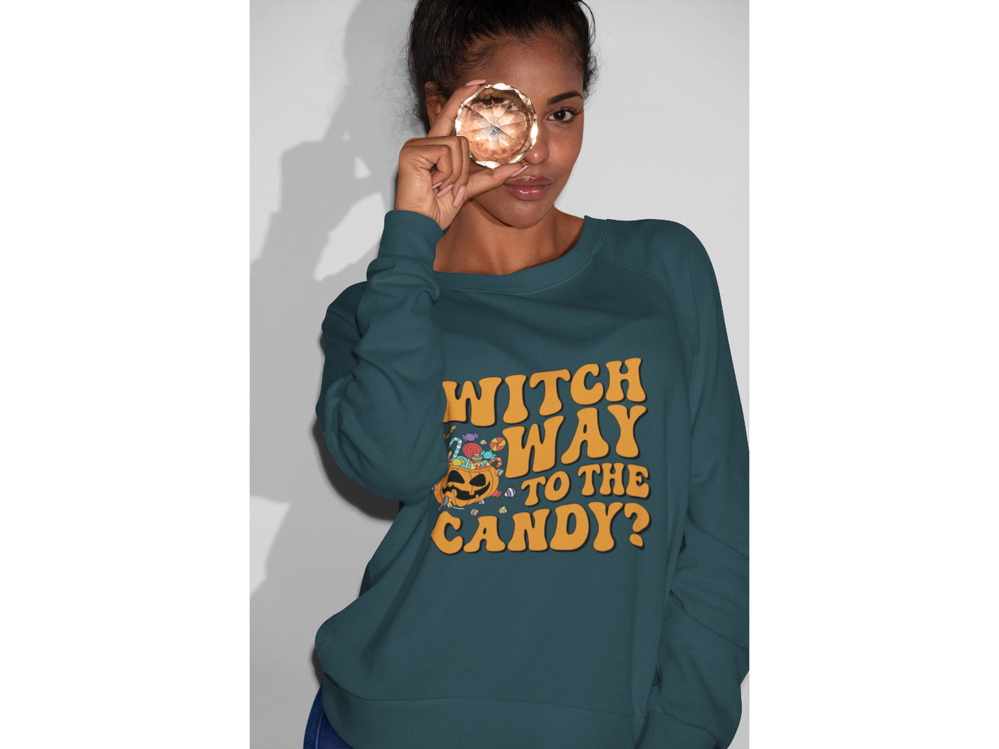 A woman wears a teal sweatshirt with "Witch Way to the Candy?" and a pumpkin graphic.