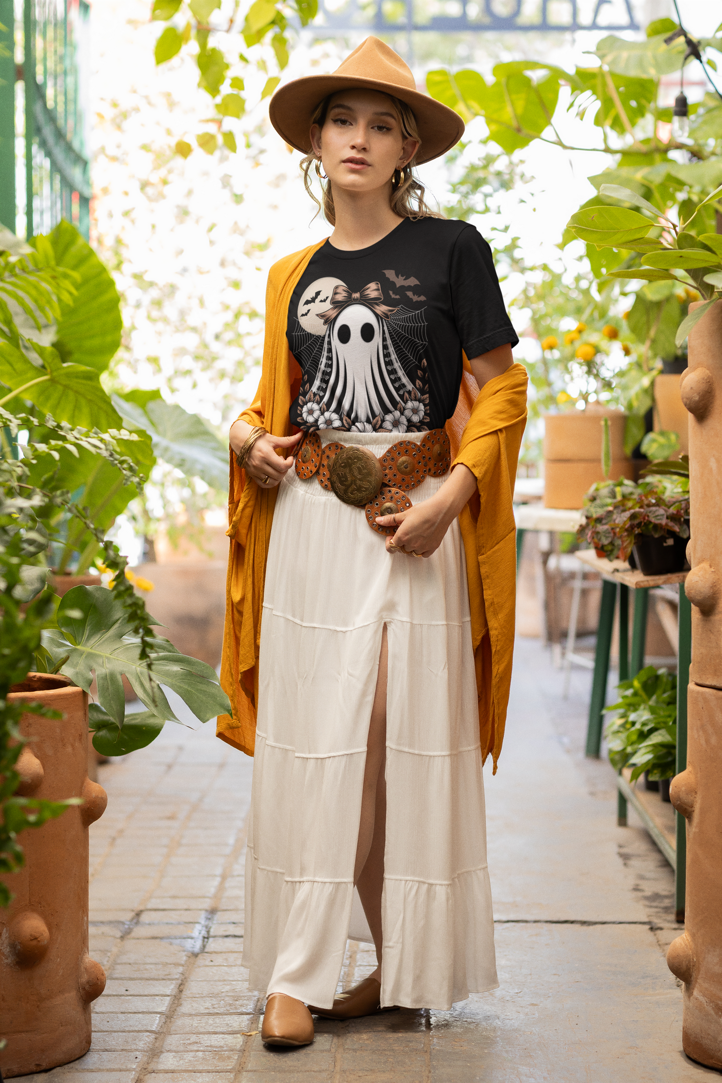 A woman wears a black t-shirt with a vintage lace ghost design, a long white skirt, and a brown hat.