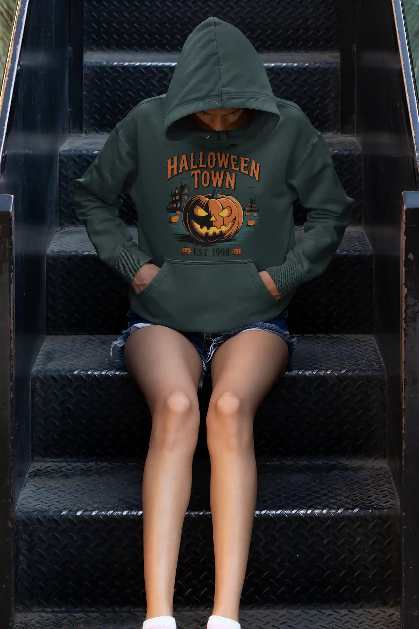A person wearing a dark green Halloween Town hoodie with a pumpkin graphic sits on metal stairs.