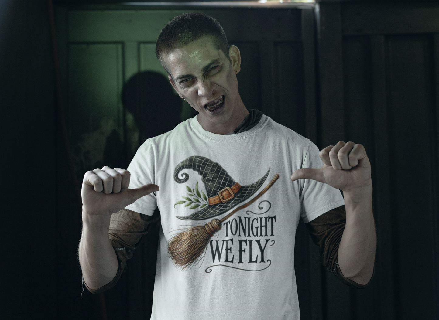 A man in zombie makeup points to a white t-shirt with a witch hat and broom.