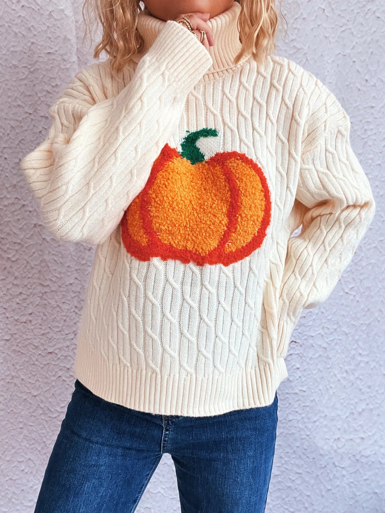 A cream cable-knit turtleneck sweater with an orange pumpkin applique.