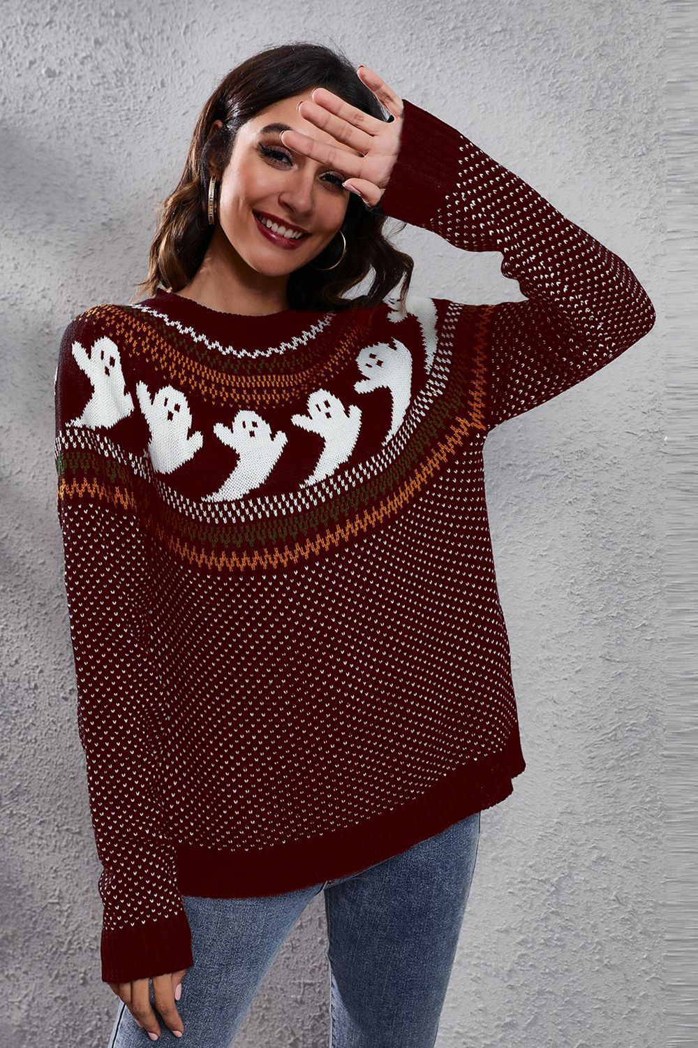 A woman smiles, shielding her eyes, wearing a maroon ghost pattern sweater and jeans.