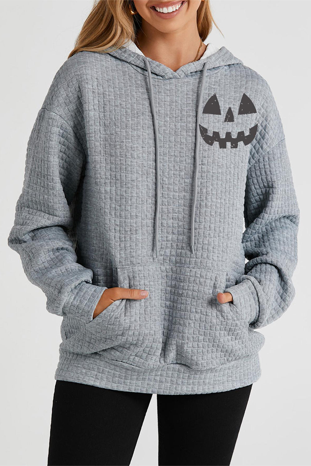 A woman wears a gray quilted hoodie with a pumpkin face graphic and front pocket.
