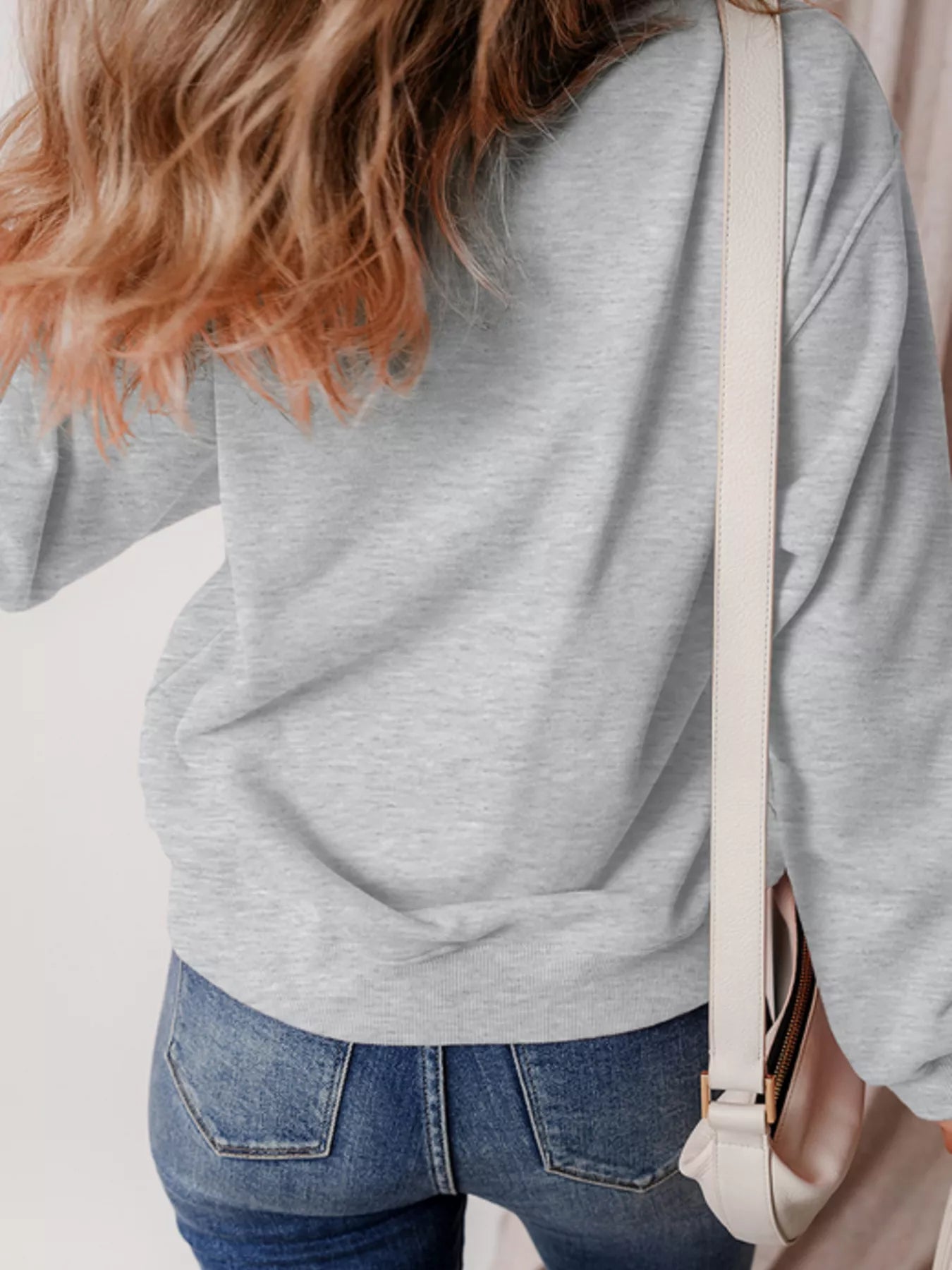 A woman from behind wears a gray sweatshirt and blue jeans, with a white bag.
