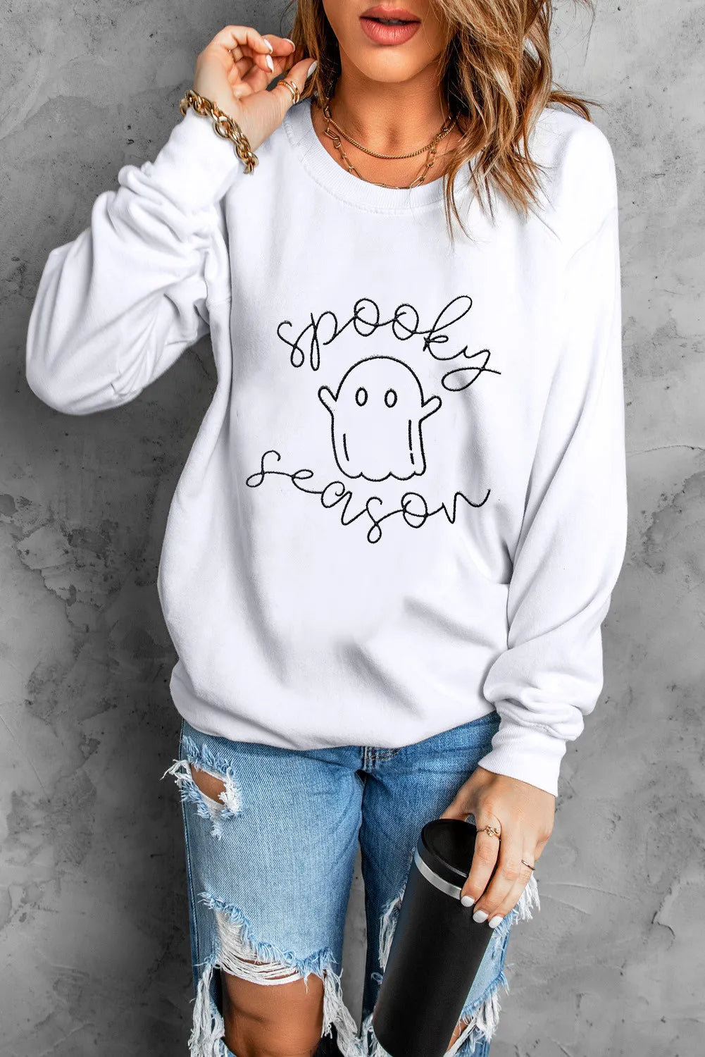 White long-sleeve sweatshirt with "spooky season" ghost graphic, worn with ripped jeans.