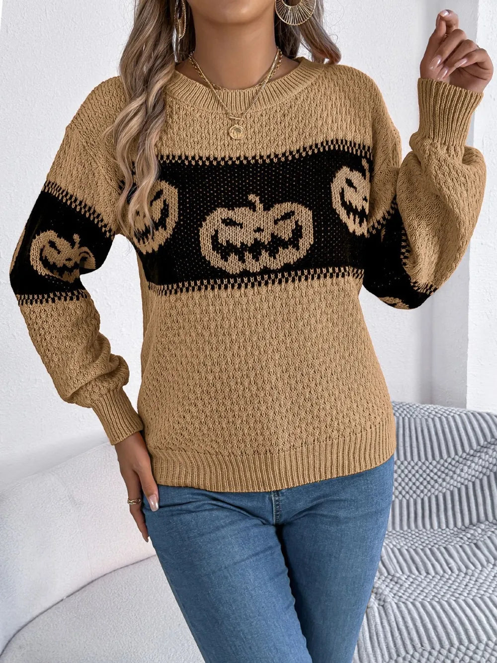 A woman wears a tan long-sleeve sweater with a black pumpkin pattern and blue jeans.