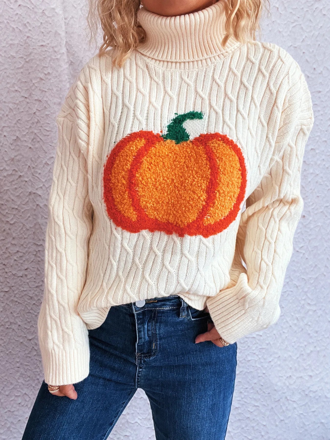 A cream cable-knit turtleneck sweater with a large orange pumpkin applique.