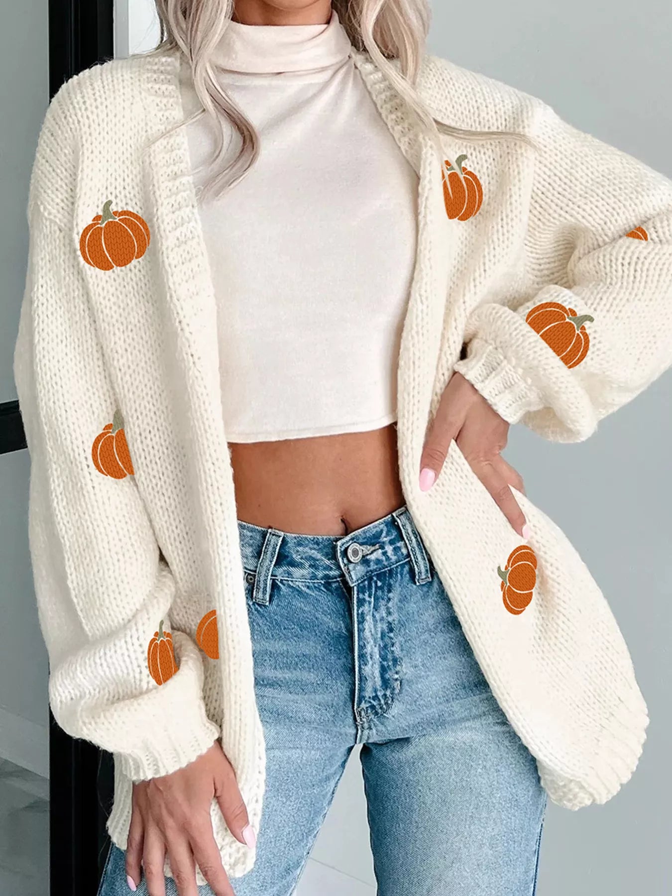 A woman wears a white knit cardigan with orange pumpkin embroidery and blue jeans.