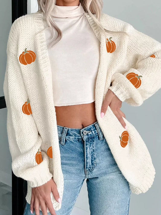A woman wears a white knit cardigan with orange pumpkin embroidery and blue jeans.