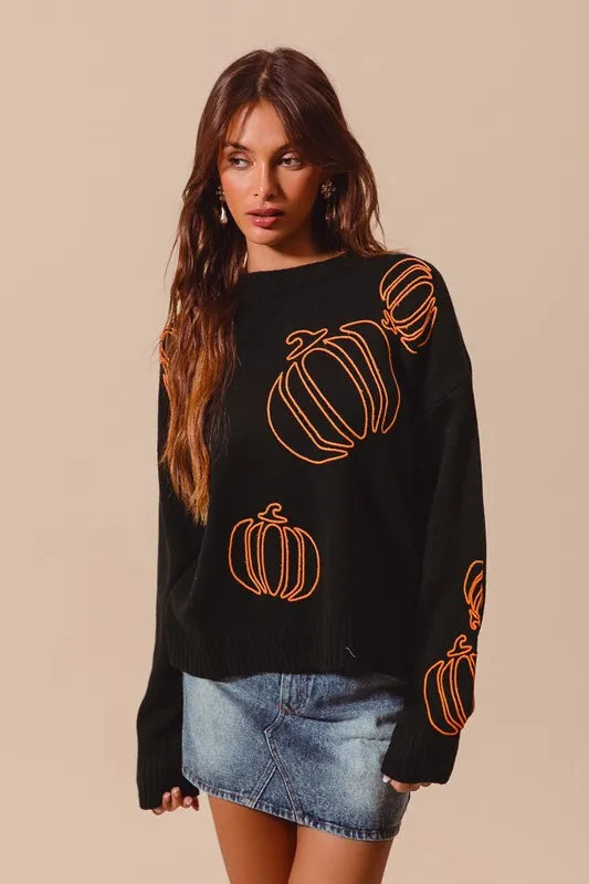 A woman models a black sweater with orange pumpkin outlines and a denim skirt.