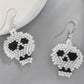 Two white and black beaded skull earrings are displayed on a gray surface.