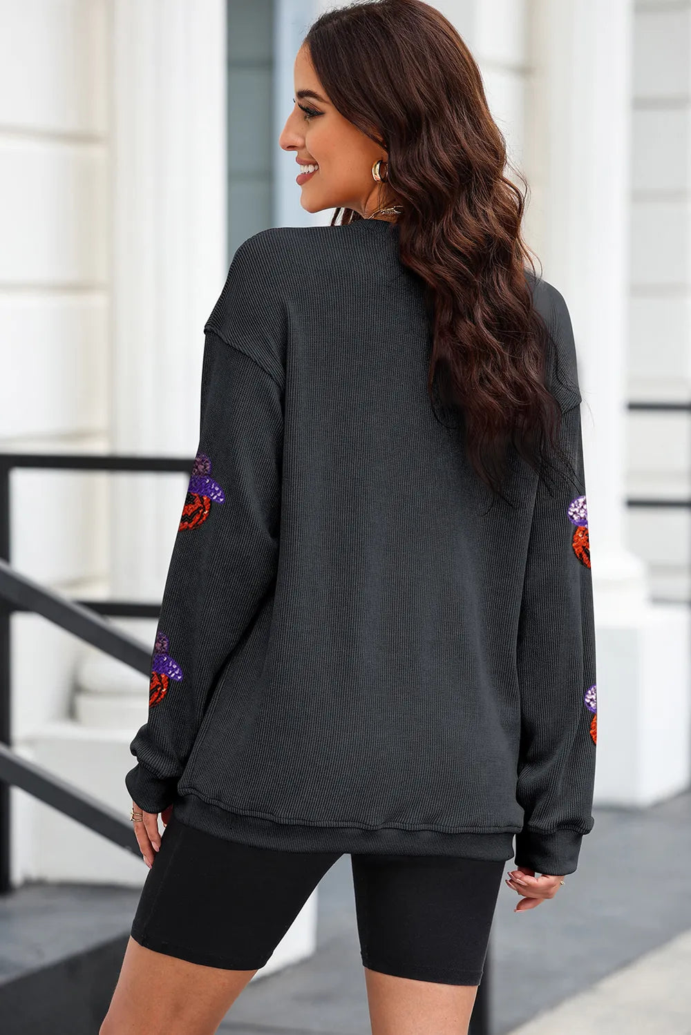 A woman from behind wears a black sequin pumpkin long sleeve sweatshirt and black shorts.