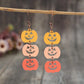 Two wooden pumpkin earrings, each with three pumpkins in yellow, pink, and orange, hang from a branch.