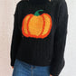 Black cable-knit turtleneck sweater with an orange pumpkin applique, worn with blue jeans.