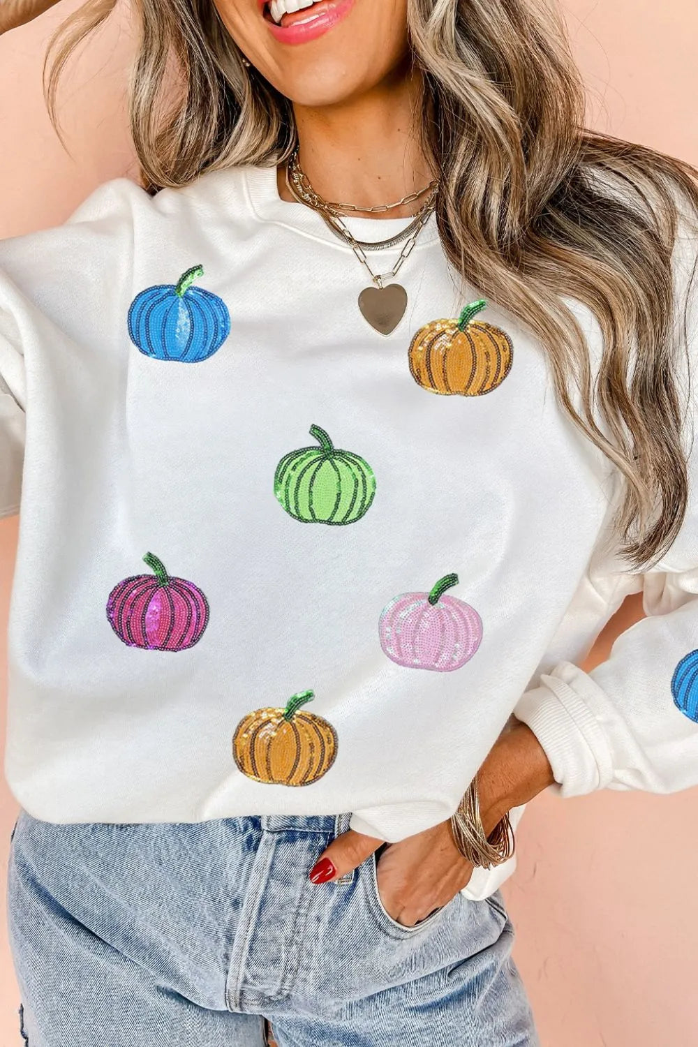 A woman wears a white long-sleeve sweatshirt with colorful sequin pumpkins.