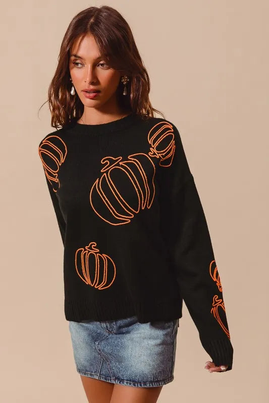 A woman models a black sweater with orange pumpkin outlines and a denim skirt.