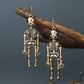 Two gold-colored skeleton alloy earrings hang from a tree branch against a dark background.