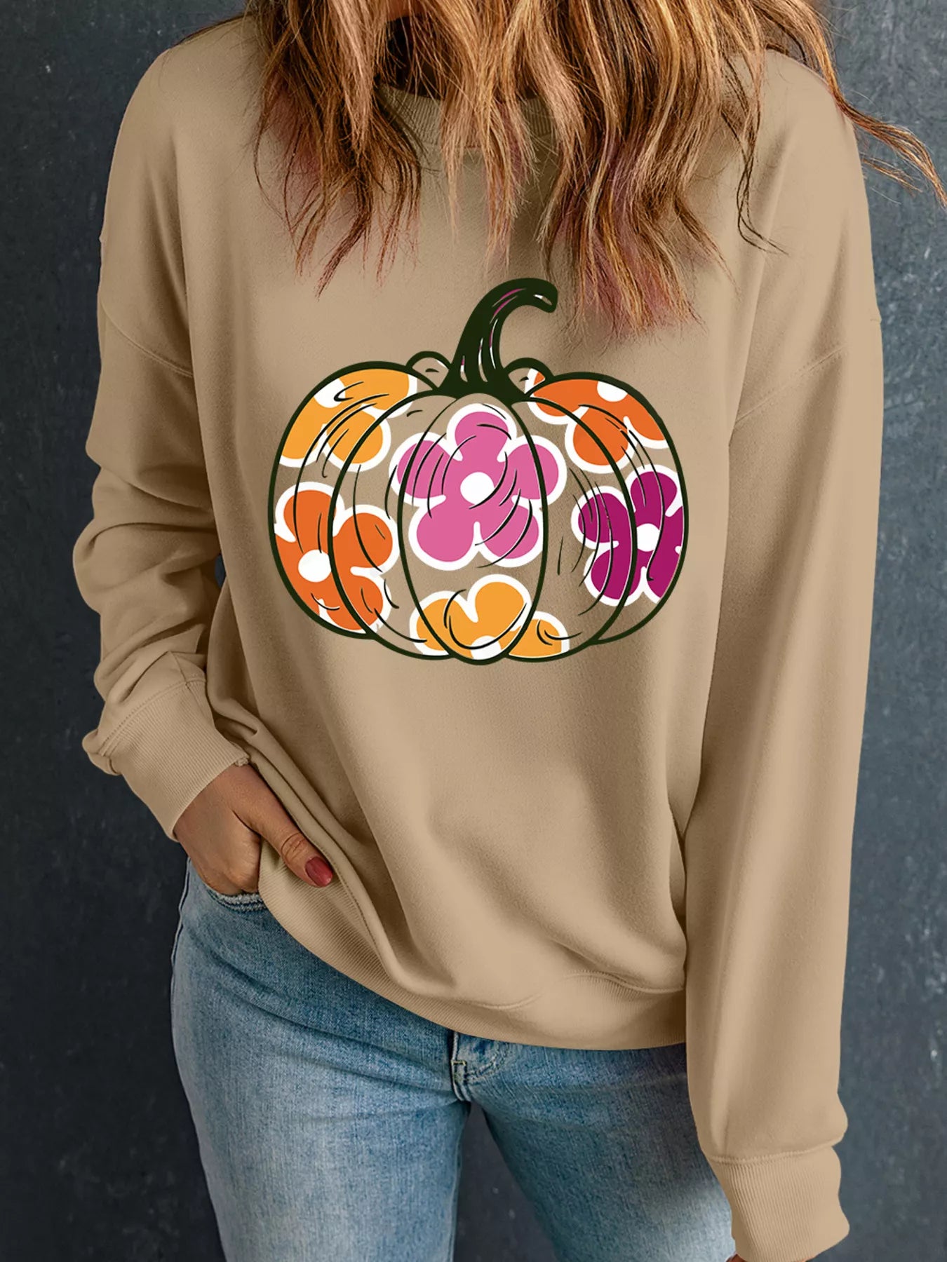 A woman wears a beige sweatshirt with a floral pumpkin graphic and blue jeans.