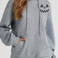 A woman wears a gray drawstring hoodie with a pumpkin face graphic and a front pocket.
