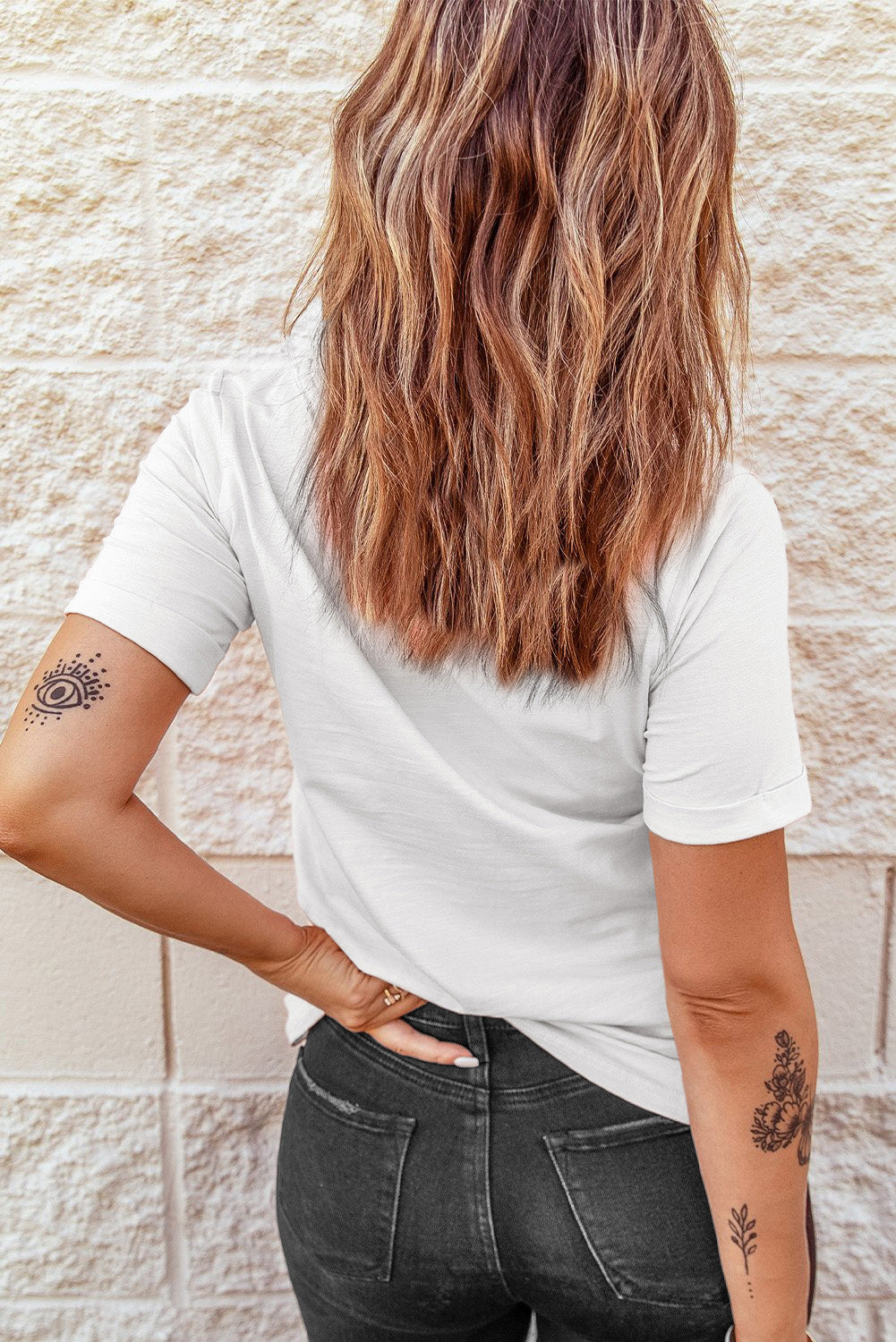 A woman with long wavy hair wears a white short-sleeve tee and dark jeans.