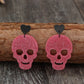 Two glittery pink skull earrings with black heart studs rest on a wooden log.