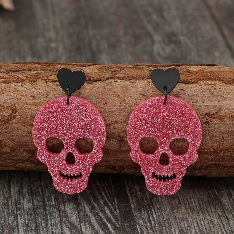 Two glittery pink skull earrings with black heart studs rest on a wooden log.