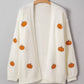 White knit cardigan with embroidered orange pumpkins, hanging on a wooden hanger.