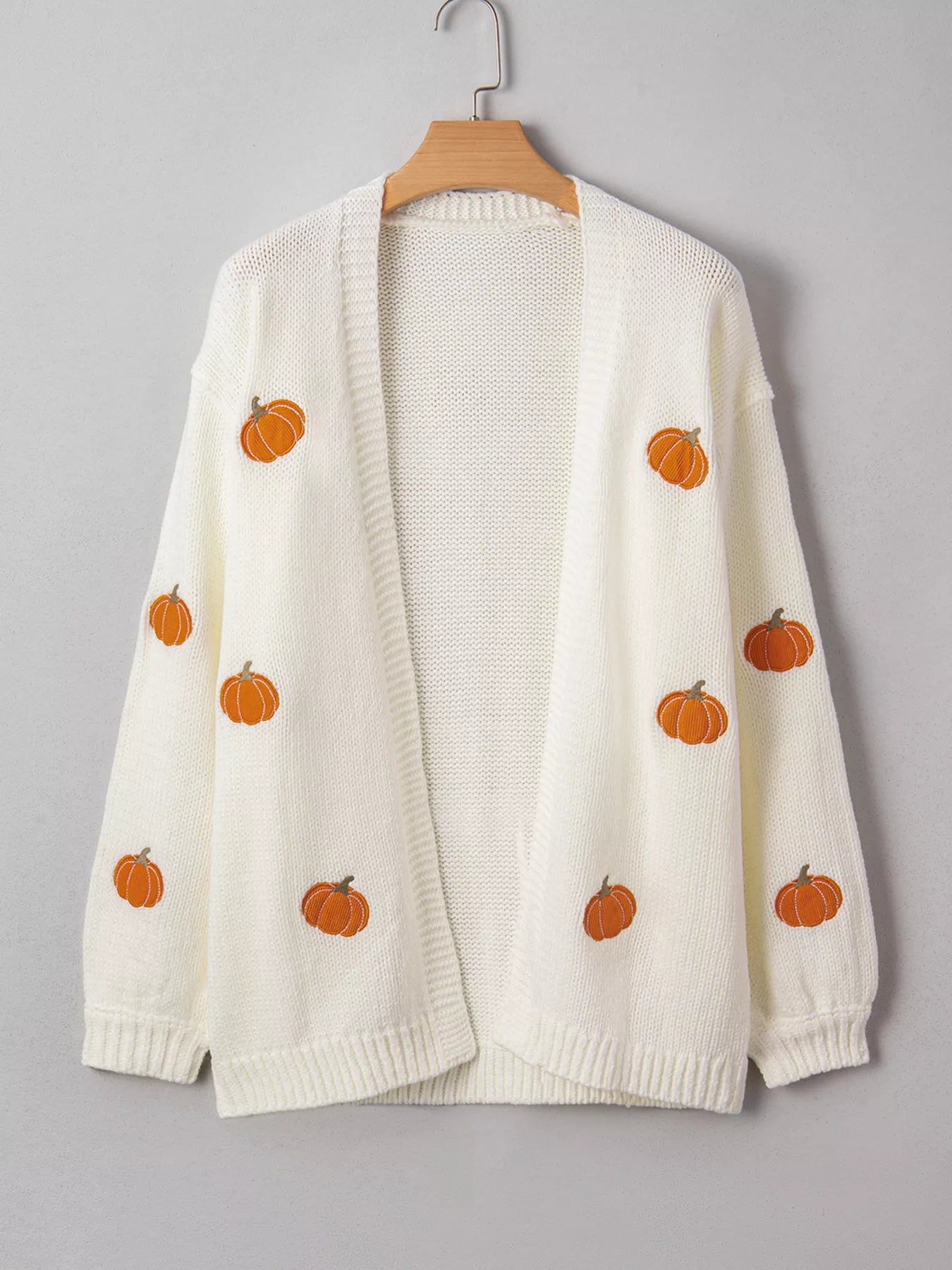 White knit cardigan with embroidered orange pumpkins, hanging on a wooden hanger.