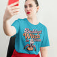 A woman in a blue t-shirt with "Baddest Witch in Town" graphic takes a selfie.
