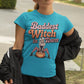 A woman wears a teal t-shirt with "Baddest Witch in Town" and a witch on a pumpkin.