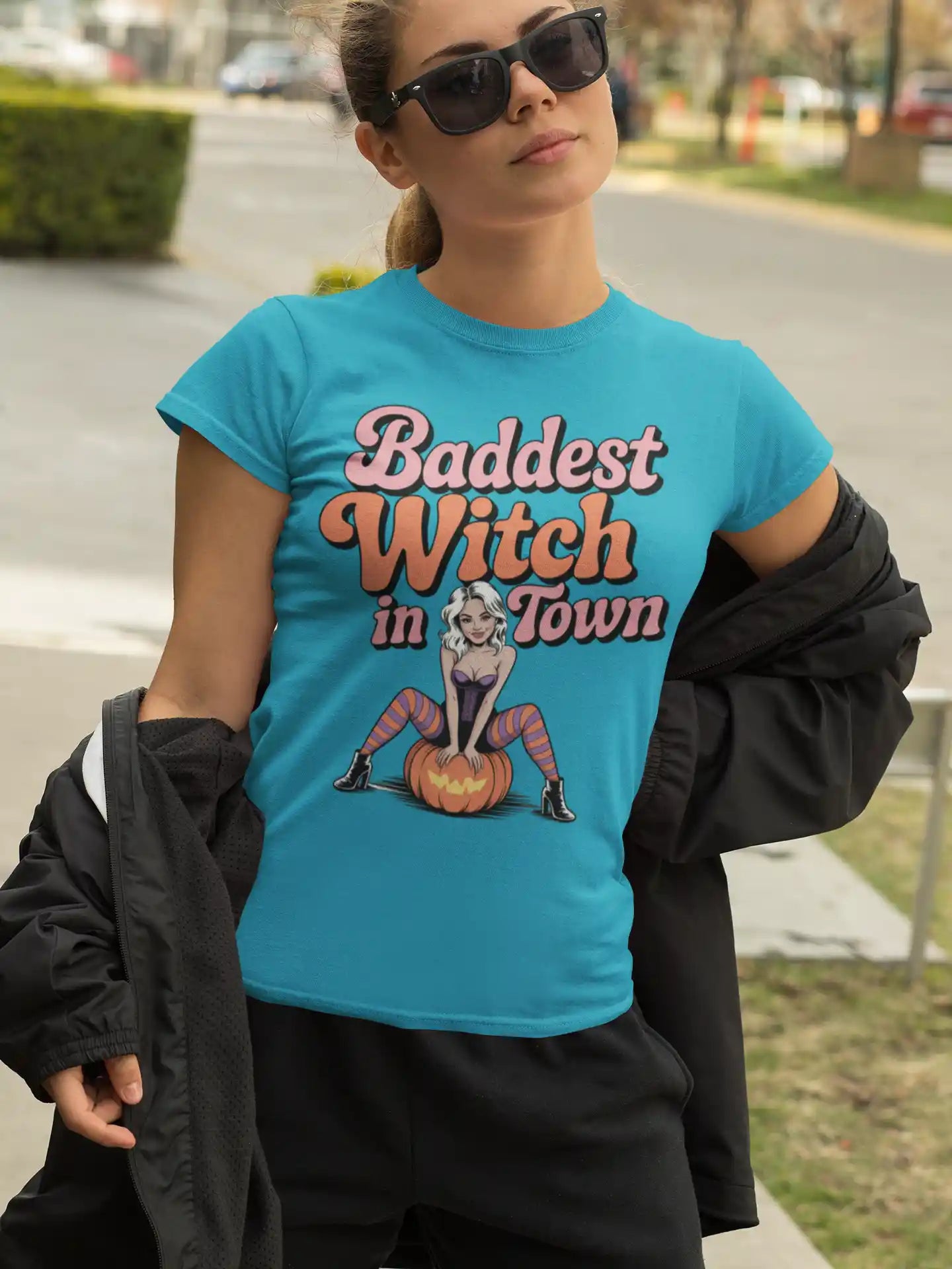 A woman wears a teal t-shirt with "Baddest Witch in Town" and a witch on a pumpkin.