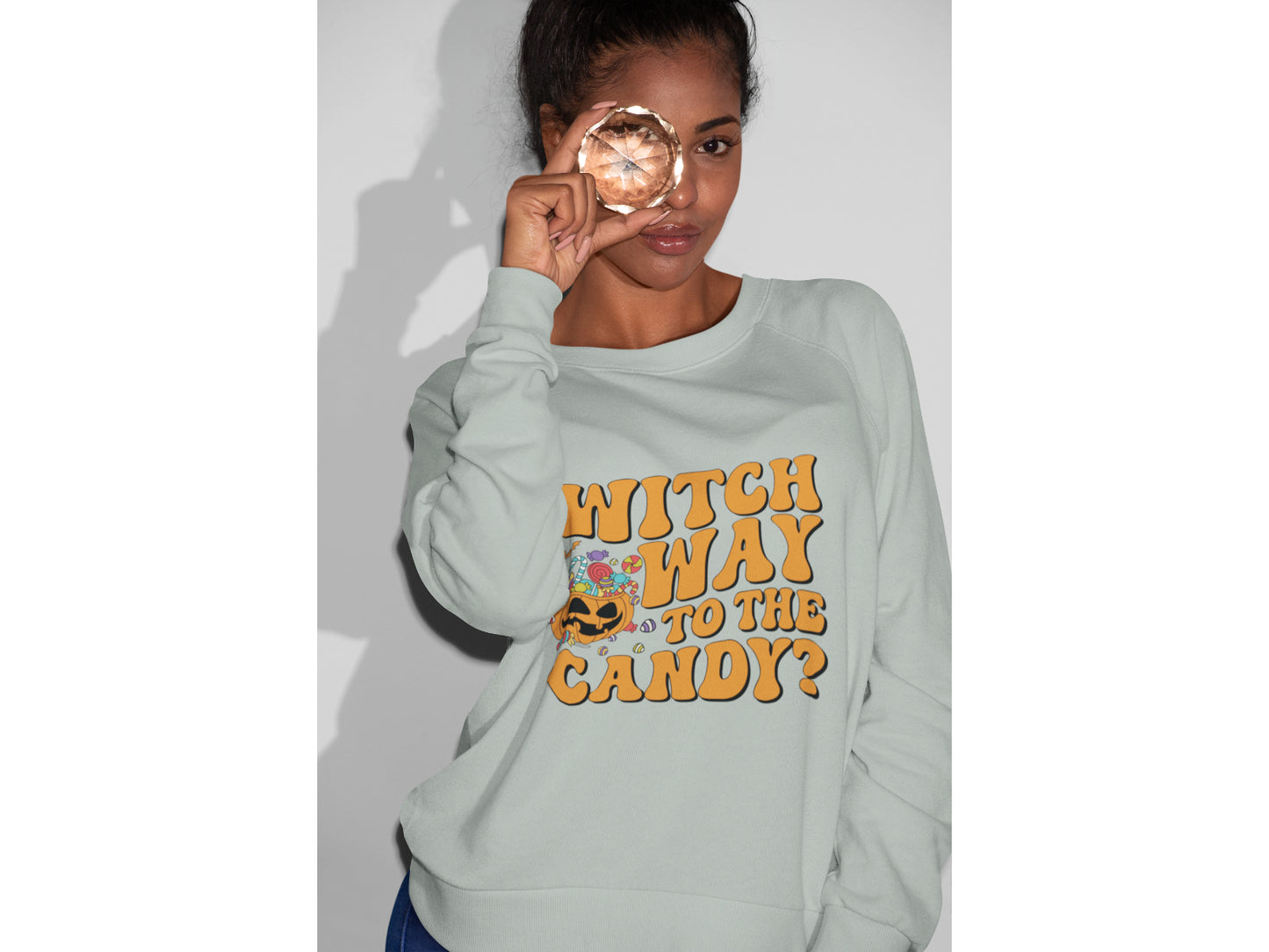 A woman wears a light gray sweatshirt with "Witch Way to the Candy?" in orange text.