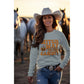 A woman in a cowboy hat and jeans wears a light blue "Witch Way to the Candy" sweatshirt.
