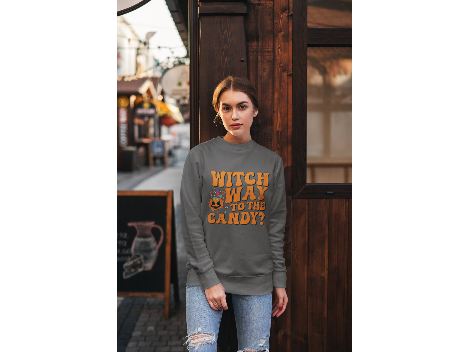 A woman wears a dark gray sweatshirt with "Witch Way to the Candy?" in orange text.