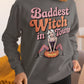 A woman wears a dark gray sweatshirt with "Baddest Witch in Town" and a witch graphic.